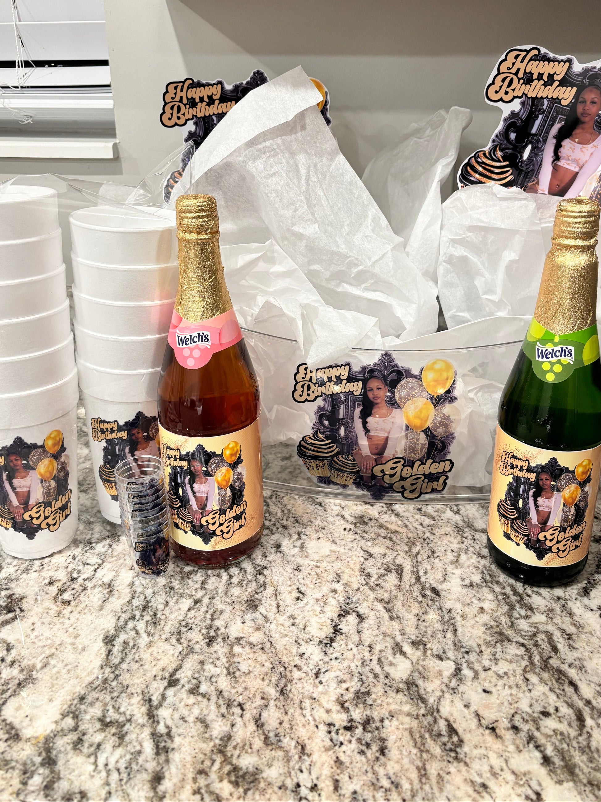 Two bottles of champagne with personalized labels on a kitchen counter, surrounded by party supplies.